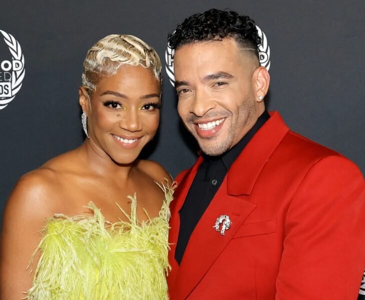 Tiffany Haddish holding a baby with Jason Lee standing beside a stroller, both smiling in an outdoor garden setting.